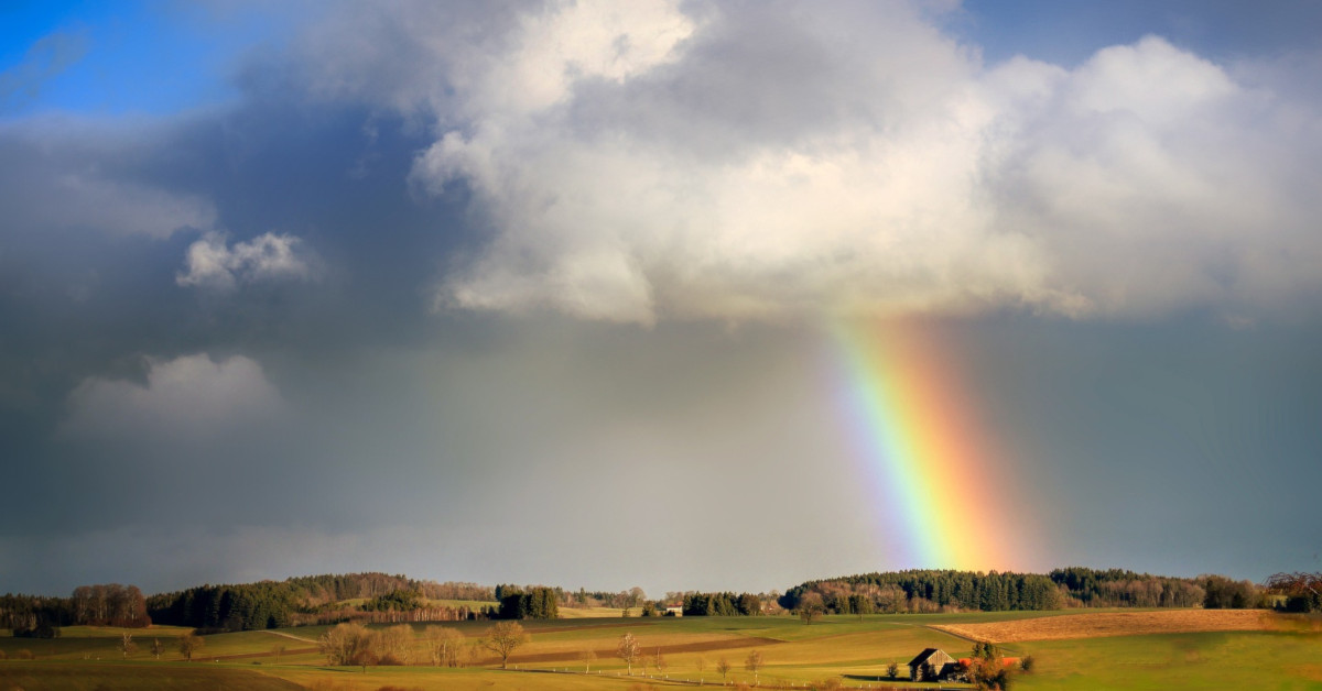 A rainbow reaches down from a storm cloud as the sun shines on a fields of gold, green, and brown, a farm house, and distant trees on hills